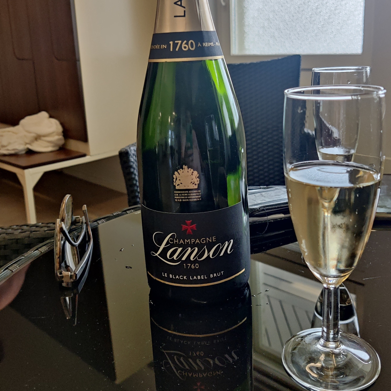 En flaske med Lanson Champagne og et fyldt champagneglas står på et sort bord, solbriller ligger ved siden af.