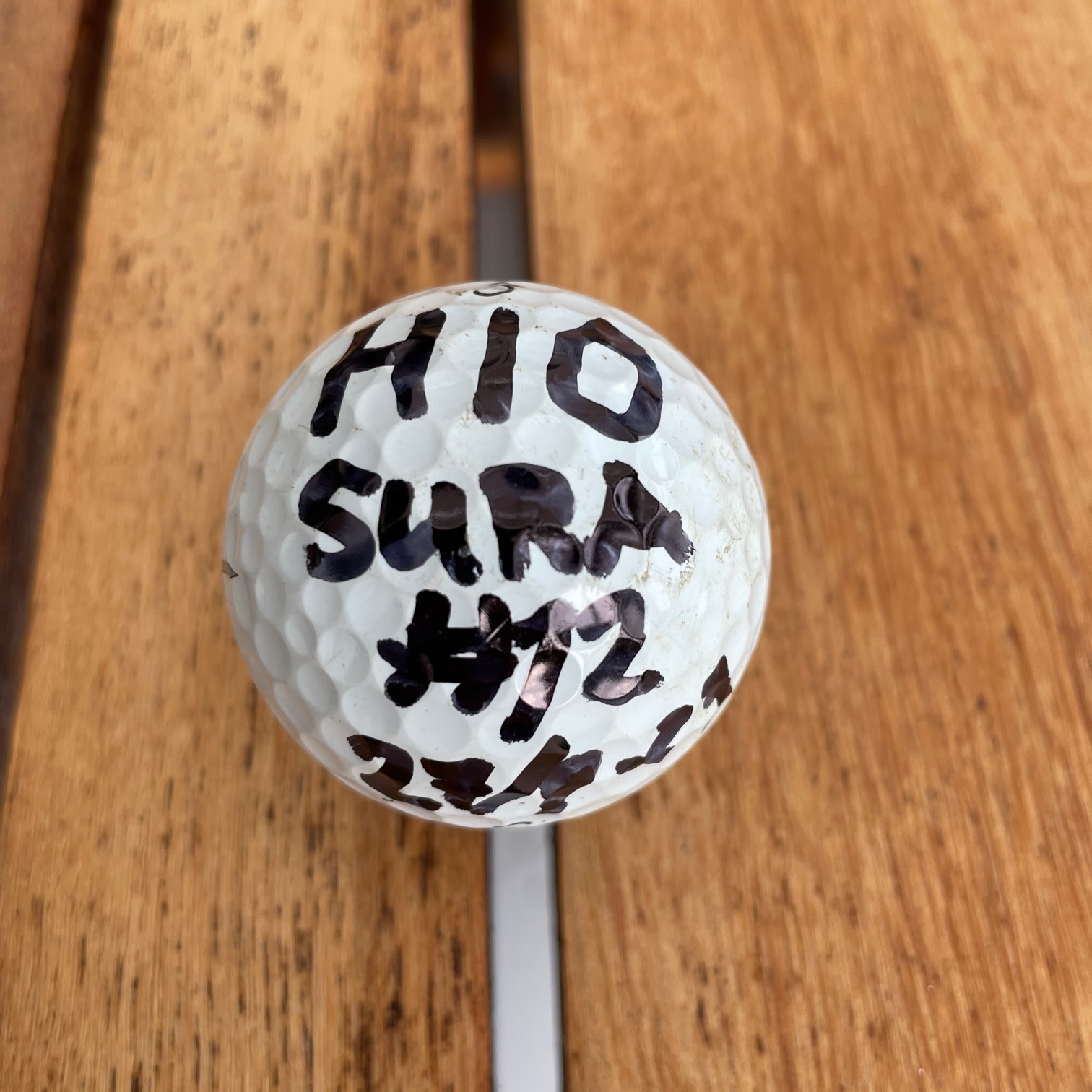 Golfboll med texten "HIO SURA #12 27/7-23" skriven med svart tusch, placerad på ett träbord.