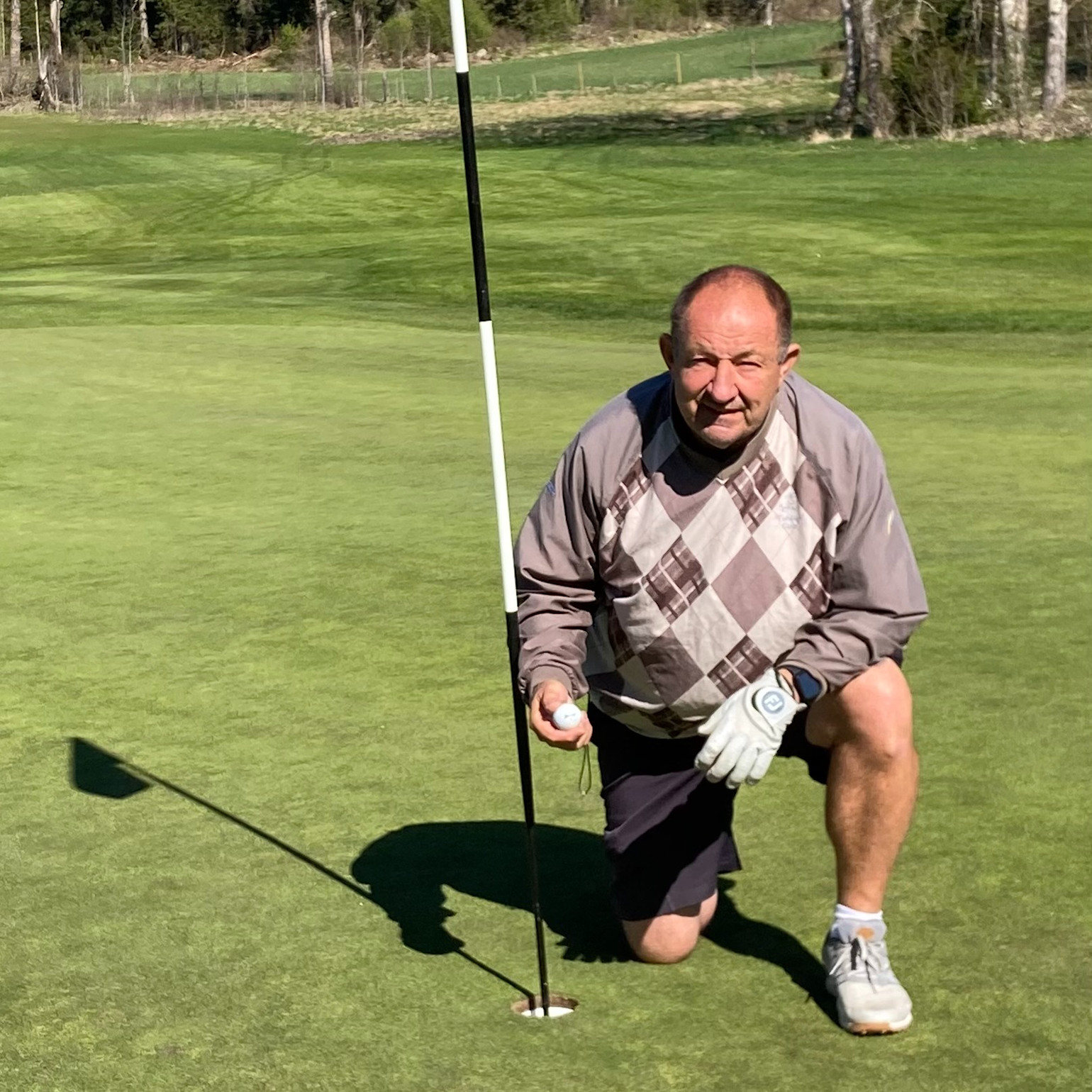 En ældre mand i golftøj knæler ved et golfhul, holder en golfbold og flagstang på en grøn golfbane.