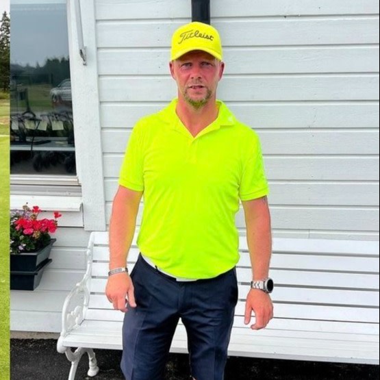 Mand med gul Titleist-kasket og gul polo står foran en hvid trævæg ved siden af en hvid bænk og en blomsterkasse.