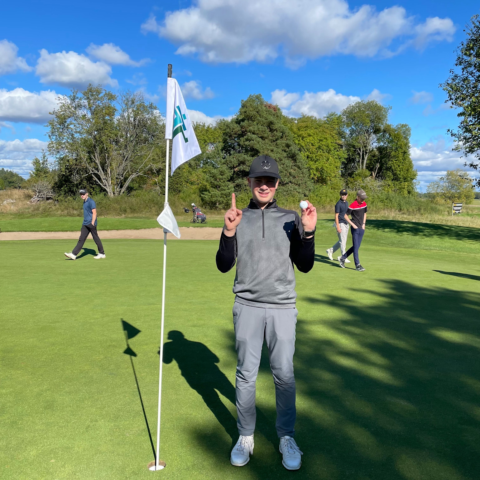 Dreng står ved siden af et golfhul og en flag, holder en golfbold op og viser én finger, solrig dag på golfbanen.