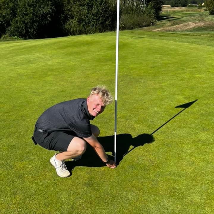 Un jeune homme en polo noir et short s’accroupit près d’un drapeau de trou de golf sur un green ensoleillé et sourit à l’appareil photo.