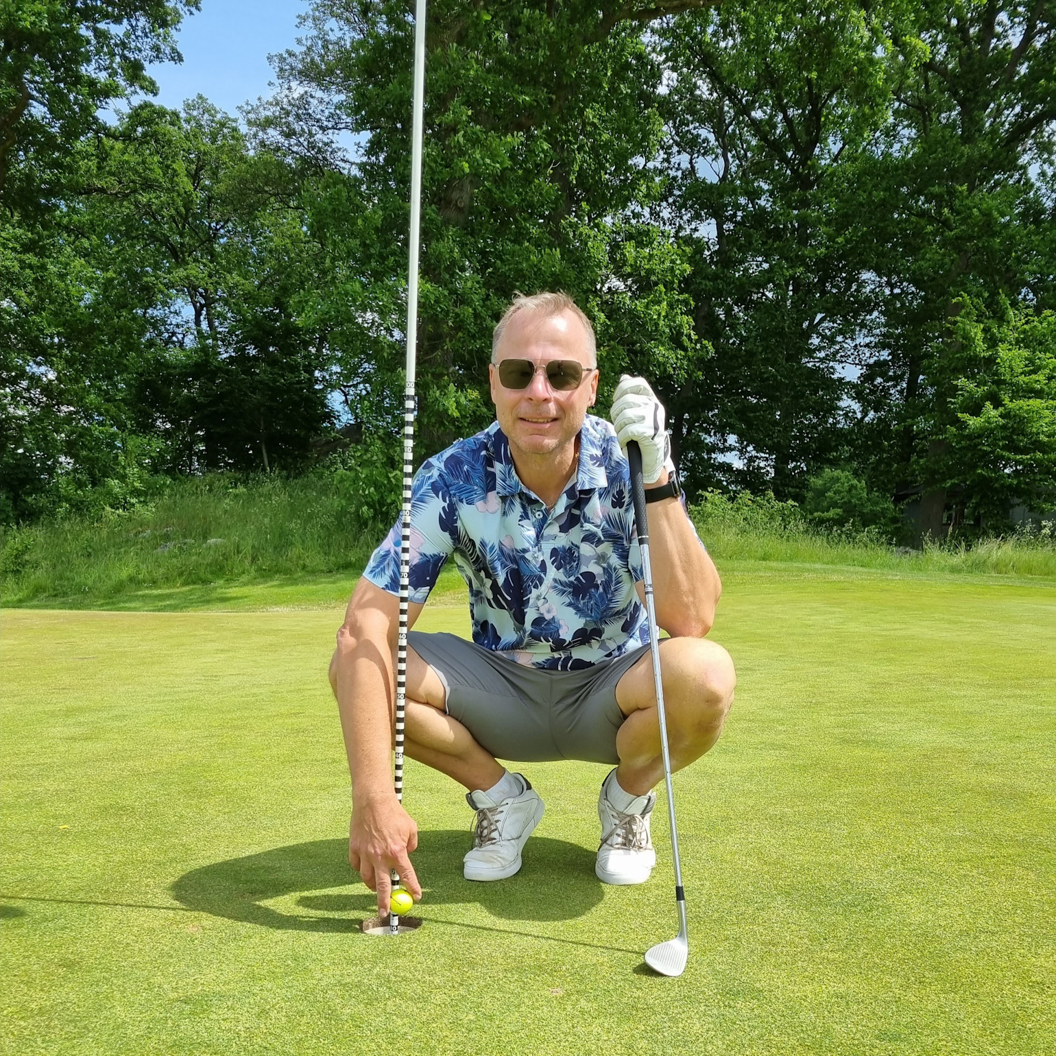 Mand i blomstret skjorte sætter sig på hug ved golfhul, holder en golfkølle og peger på en golfbold på greenen, omgivet af grønne omgivelser.