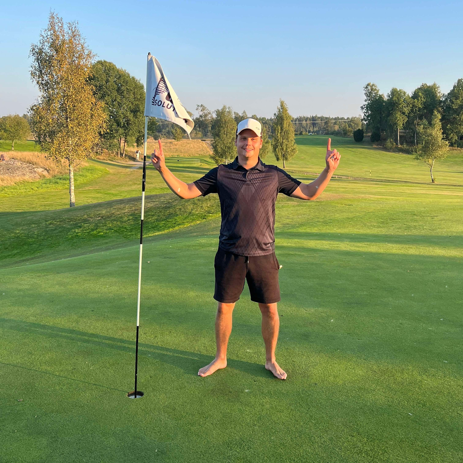 Barfota man i svart pikétröja och shorts står bredvid en golfflagga på en grön golfbana och pekar uppåt med båda händerna.