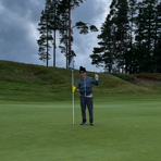Person står på en golfbana och håller i flaggstången på greenen, omgiven av gräs och tallar under molnig himmel.