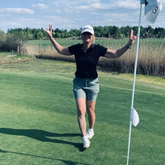 Kvinna i golfkläder gör segertecken med båda händerna bredvid en flaggstång på en golfbana under solig dag.