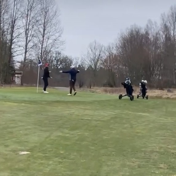 Två golfare firar vid ett hål på en golfbana, med golfbagar på vagnar bredvid och träd i bakgrunden.