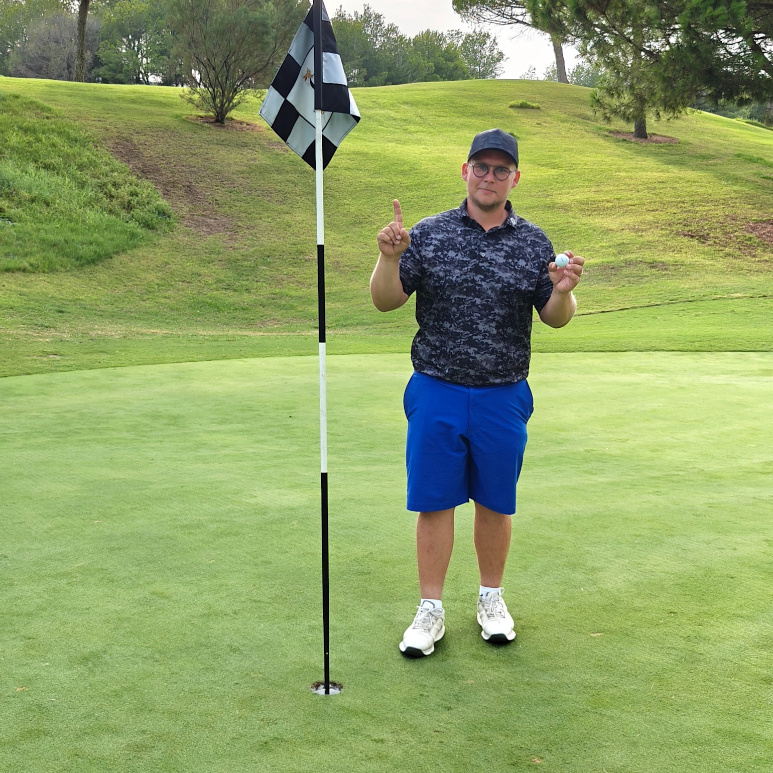 Someone is holding up a golf ball and pointing with a finger next to a flagpole on a golf green surrounded by grass and trees.