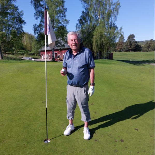 An elderly man stands next to a golf hole with a flag, holding a golf ball and smiling on a green golf course with trees and a red cottage in the background.