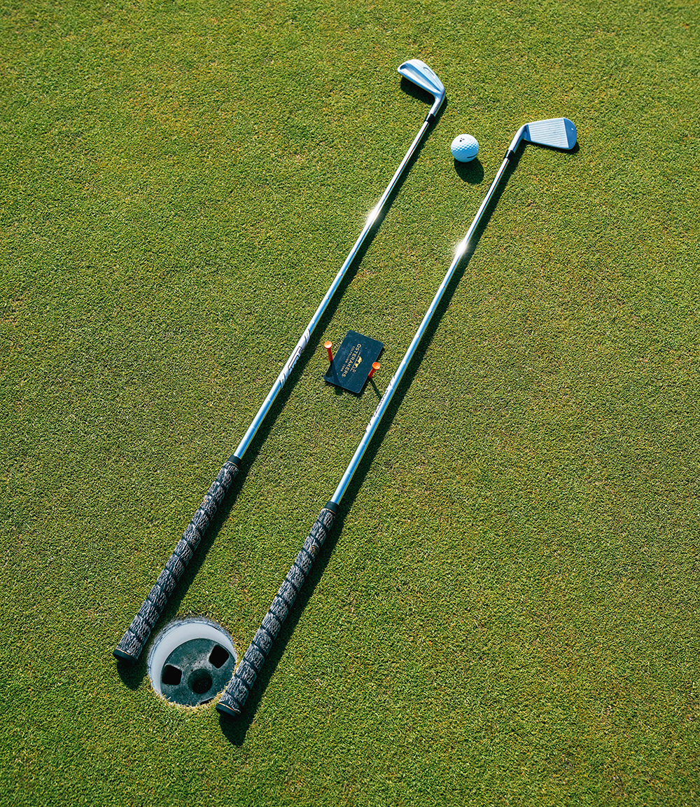 Två golfklubbor ligger parallellt på en green bredvid ett golfhål, en golfboll och ett litet svart kort med text.