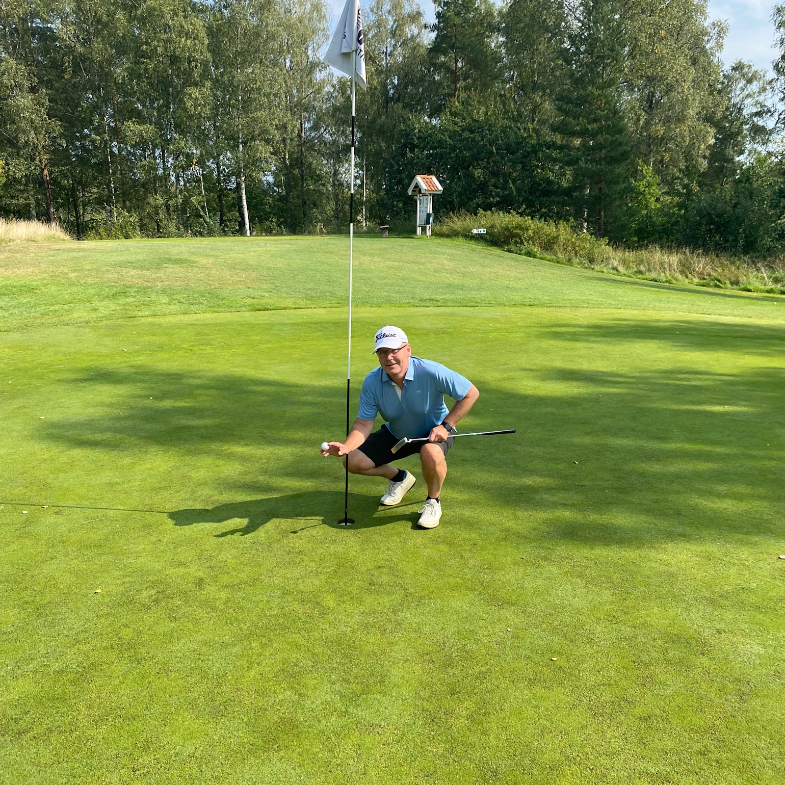 Man i blå pikétröja och vit keps hukar vid golfhål, håller golfboll och klubba på en grön golfbana med träd i bakgrunden.