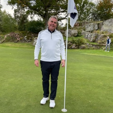 Mand i hvid golftrøje står smilende ved siden af en flagstang på en golfbane med græs, træer og klipper i baggrunden.