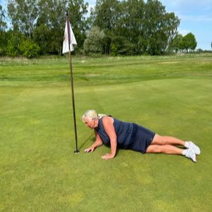 Kvinna i sportkläder ligger på gräset vid en golfbana och tittar ner i golfhålet bredvid flaggstången.