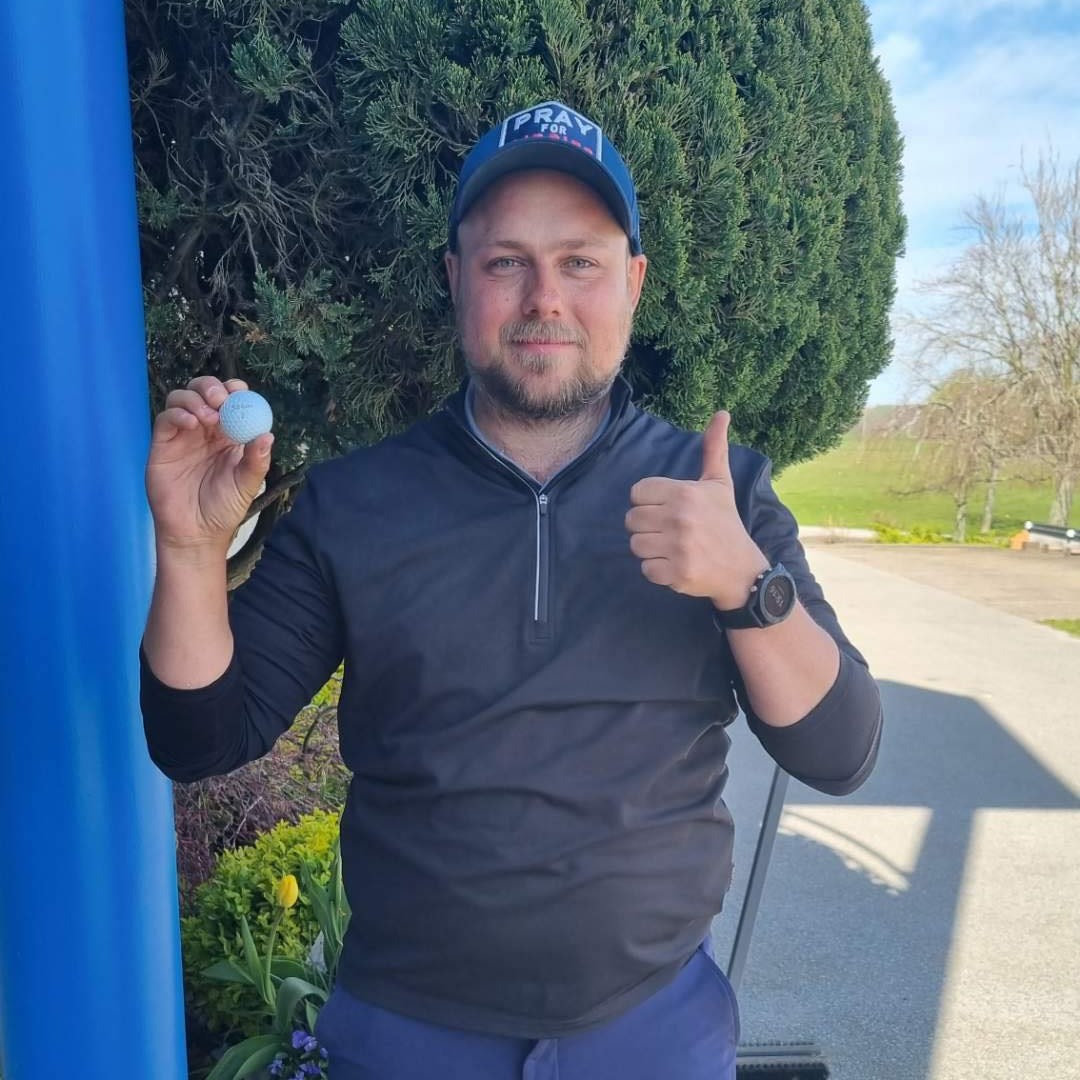 Man holder en golfbold i den ene hånd og giver thumbs up med den anden, står udendørs ved en busk.