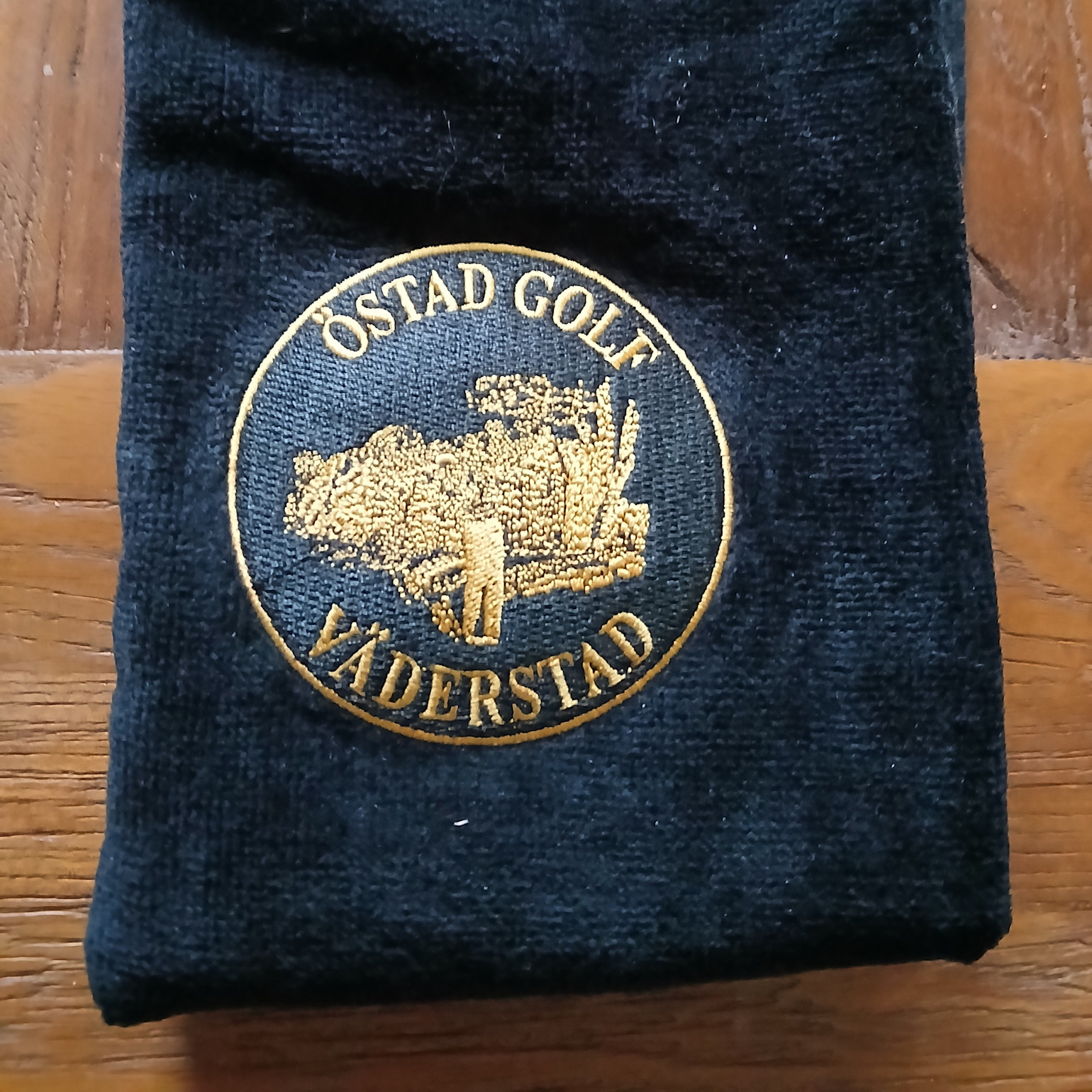 Musta kangas, jossa on kirjailtu keltainen merkki, jossa lukee "Östad Golf Väderstad" ja jossa on golfari puiden edessä.