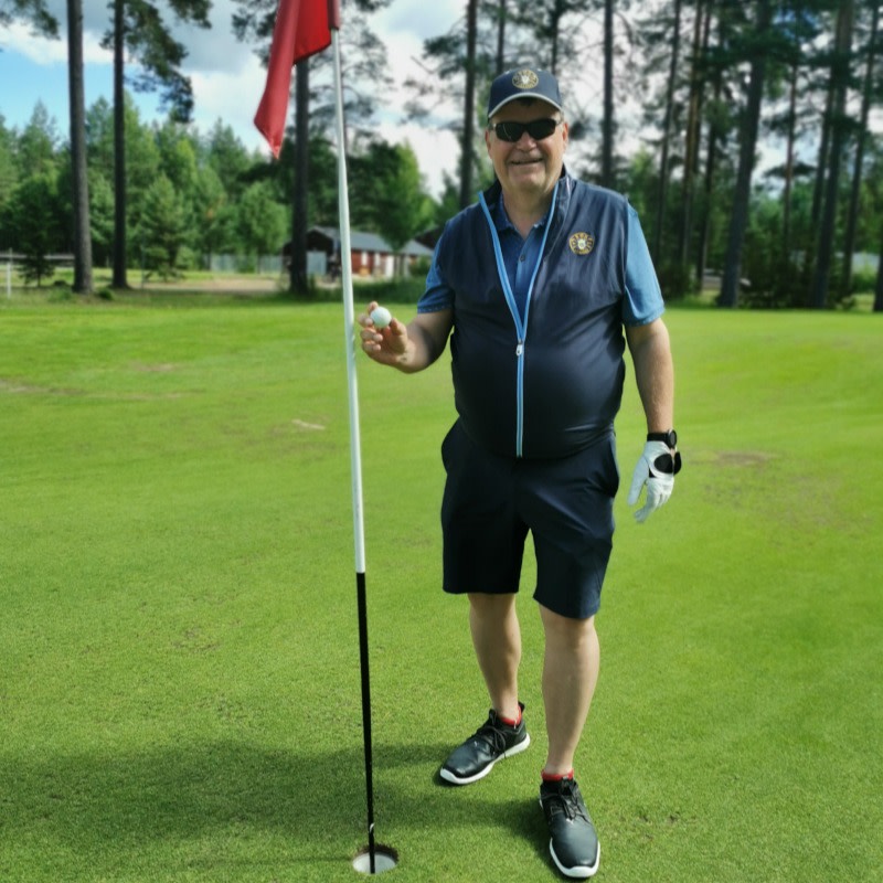 En mand i golfbeklædning holder en golfbold ved siden af hullet på en grøn golfbane med træer i baggrunden.