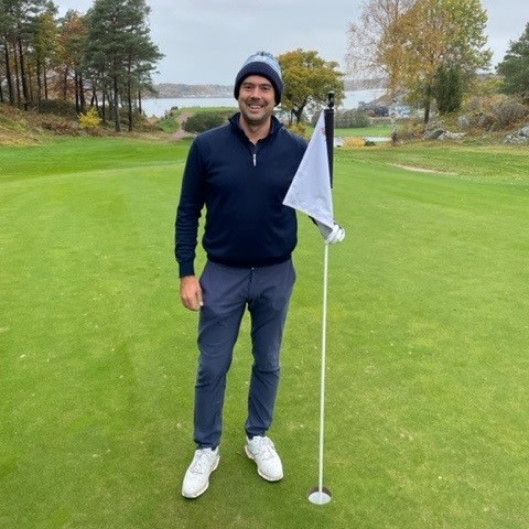 Man står på en golfbana och håller i en flaggstång på greenen, omgiven av träd och sjö i bakgrunden.