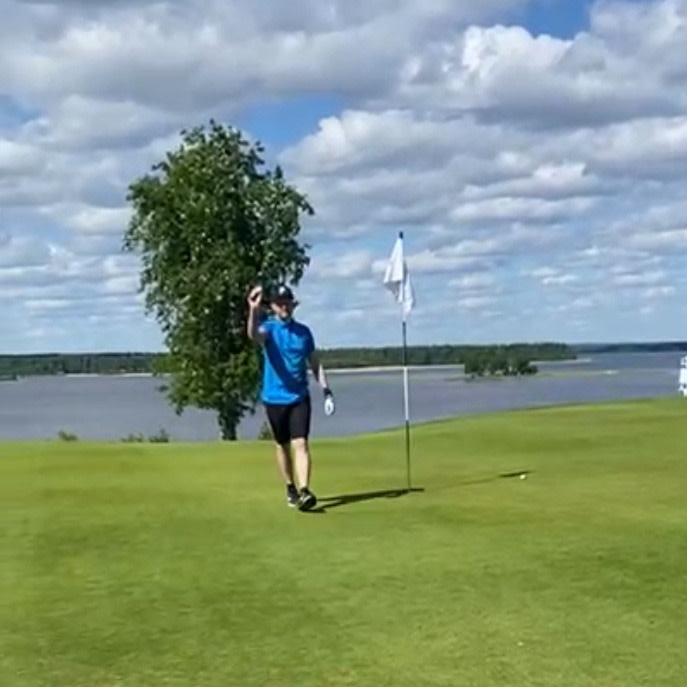 Golfspiller i blå trøje vinker ved siden af et hul med flag på en grøn golfbane, sø og træer i baggrunden.