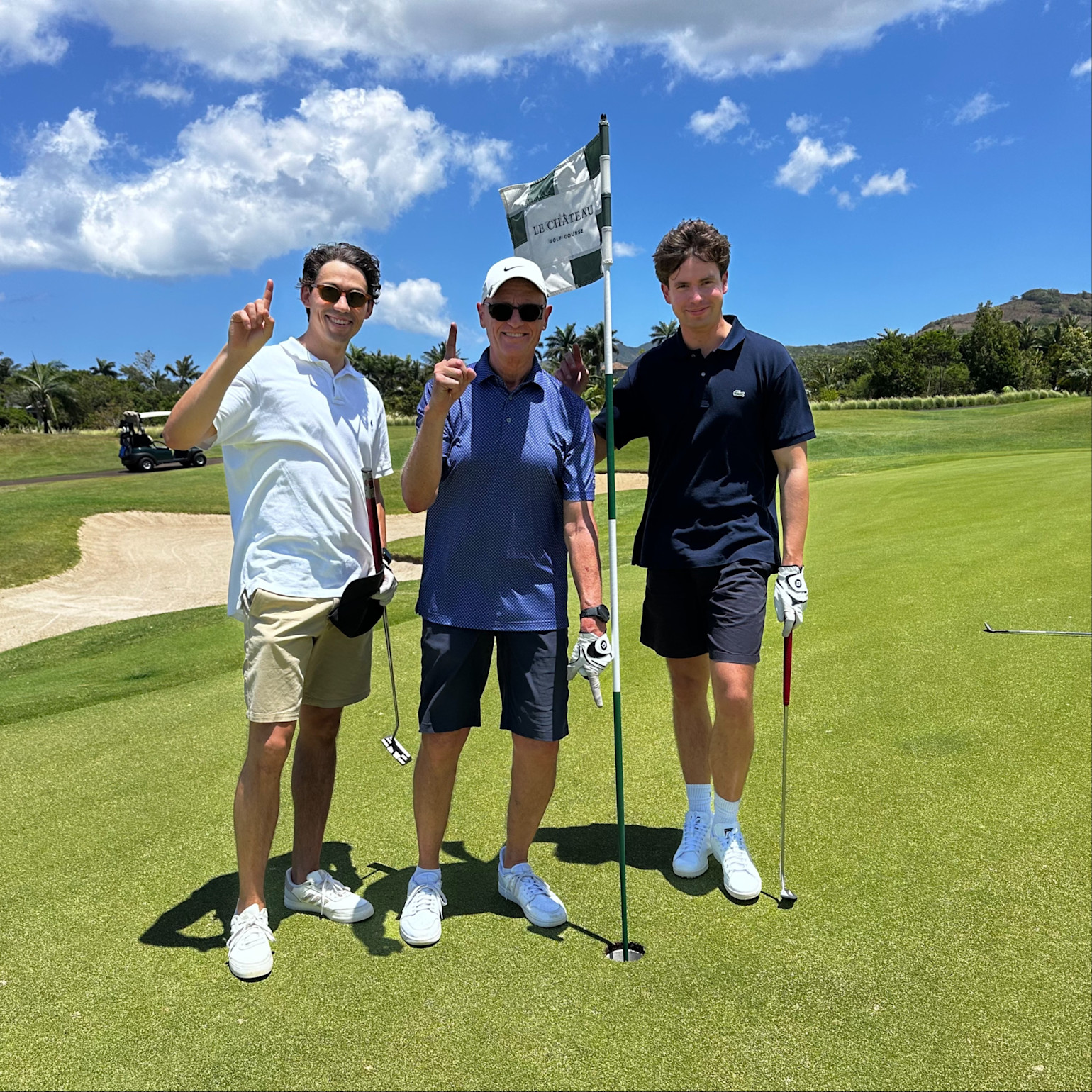 Tre män står vid ett golfhål och håller upp ett finger, med golfklubbor i händerna på en solig golfbana.