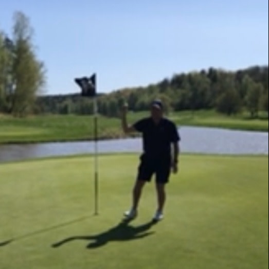 Person står på en golfgreen bredvid flaggstången och vinkar, med en damm och träd i bakgrunden under klar himmel.