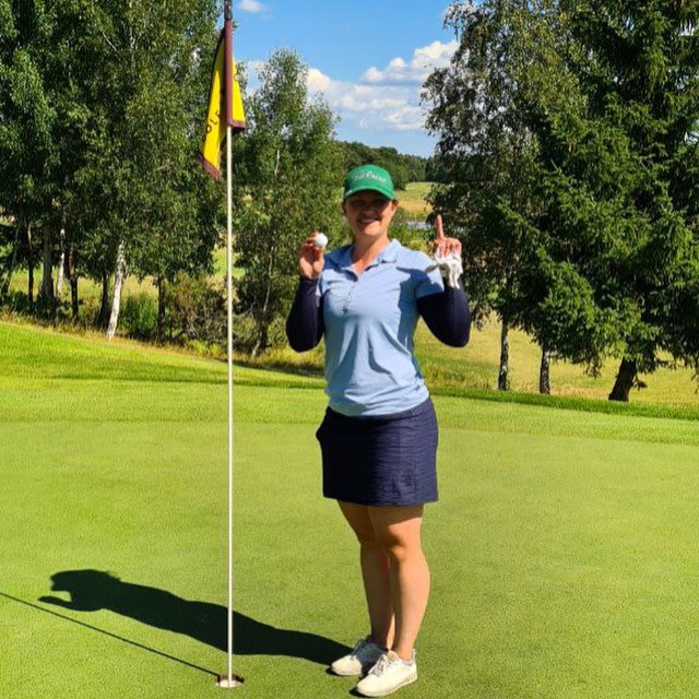 Kvinde står på en golfgreen ved siden af flagstangen, holder en golfbold op og viser én finger, omgivet af træer og blå himmel.