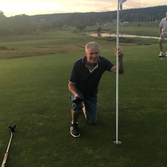 Man knæler ved siden af golfhullet og holder i flagstangen på greenen, en golfkølle ligger på jorden, solnedgang i baggrunden.