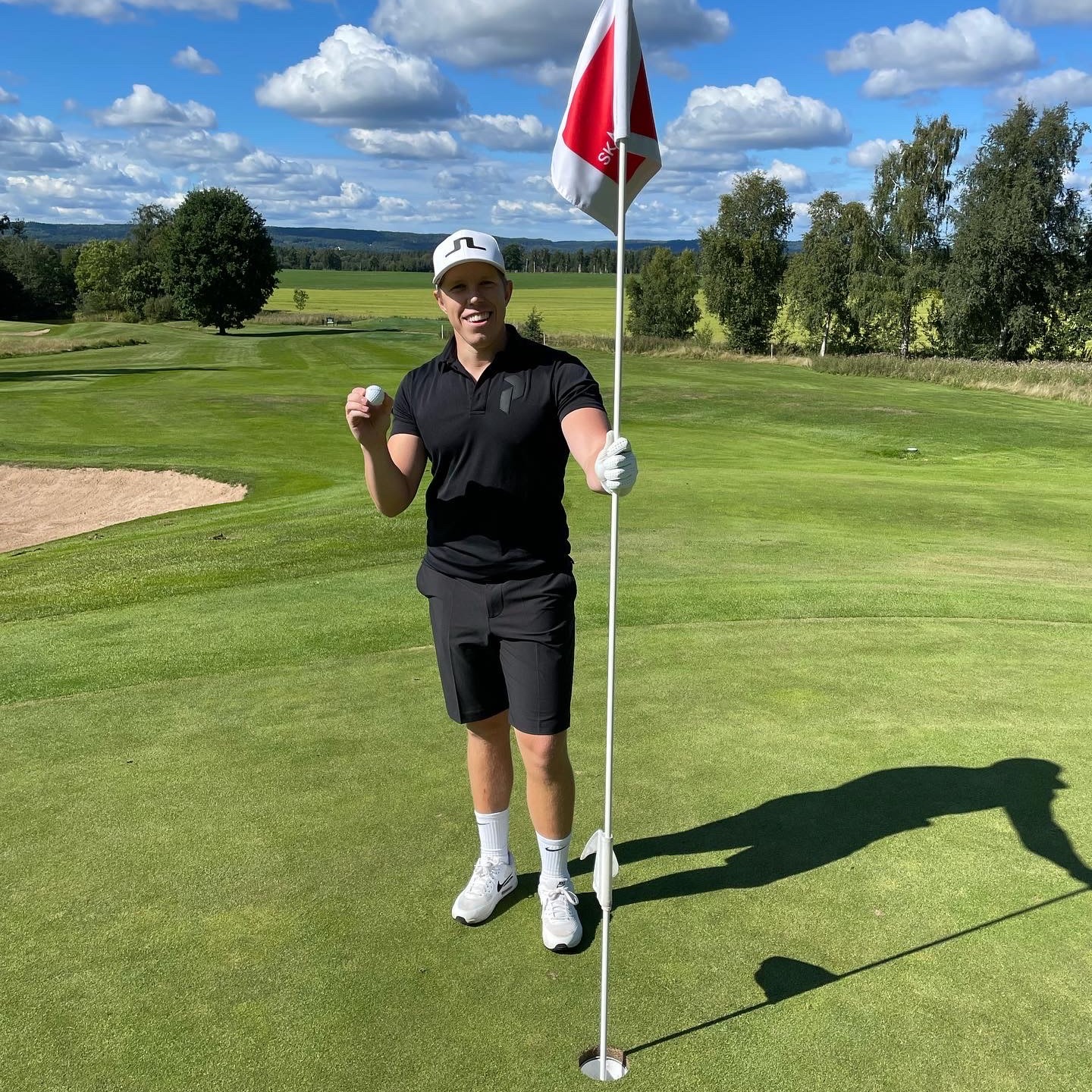 En person holder en golfbold og en flagstang ved et golfhul på en grøn golfbane under en solrig himmel.
