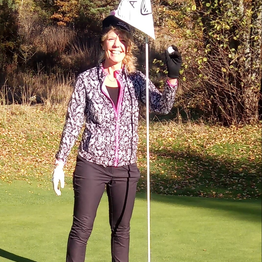 Kvinde står smilende på en golfbane, holder en golfbold i den ene hånd og flagstangen i den anden, omgivet af efterårsblade.