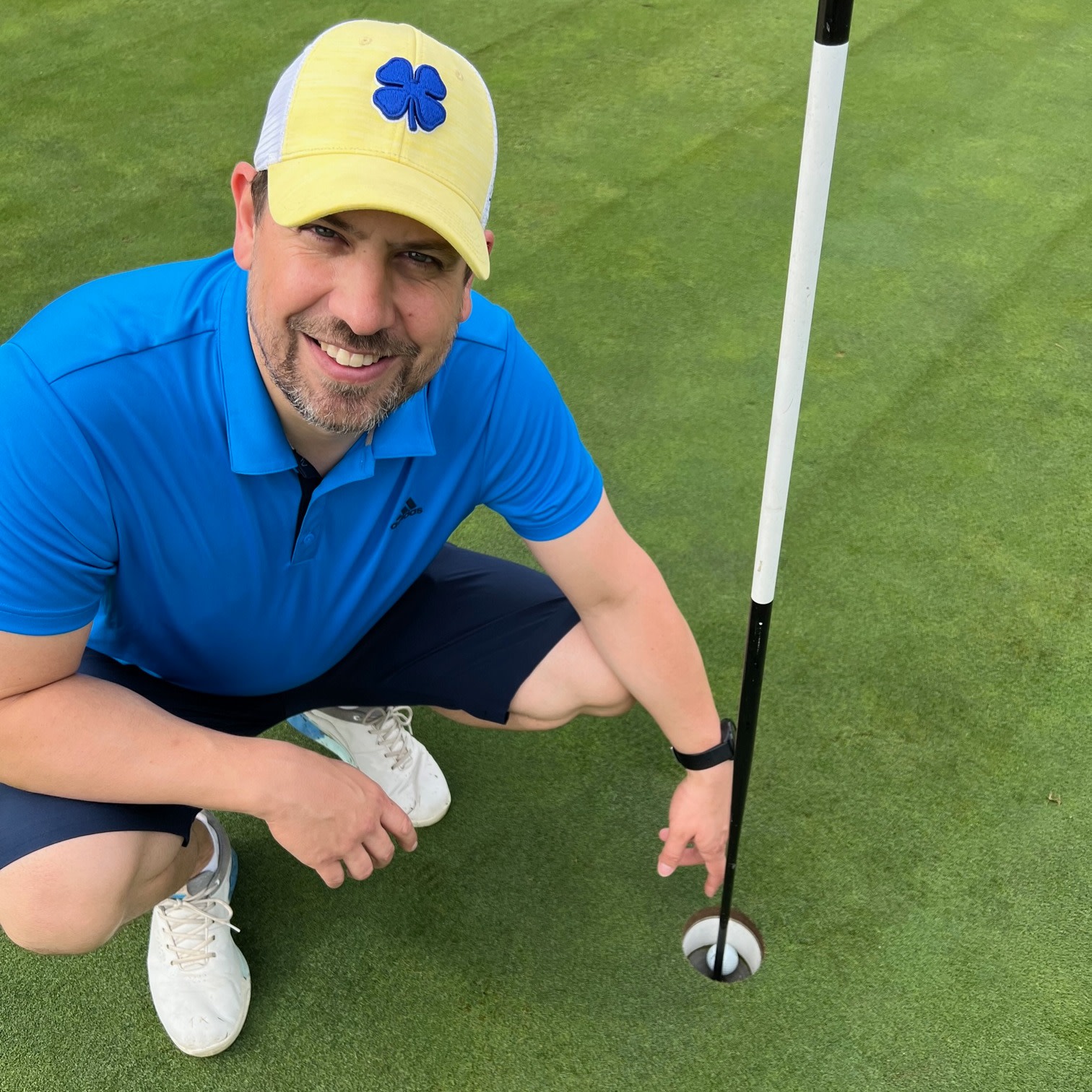 Mand i blå polo og gul kasket peger smilende på en golfbold i hullet på en golfbane.