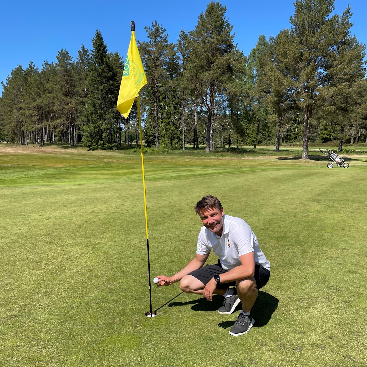 Mand i hvid polo-trøje hukker ved golfhul og holder en golfbold ved siden af flagstangen på en grøn golfbane med træer i baggrunden.