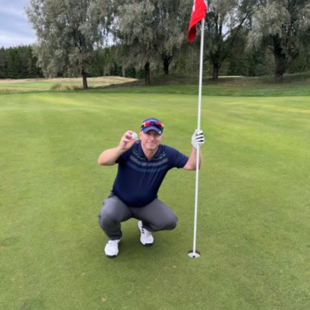 Mand i golfbeklædning hukker ved et golfhul, holder en golfbold og flagstang på en grøn golfbane med træer i baggrunden.