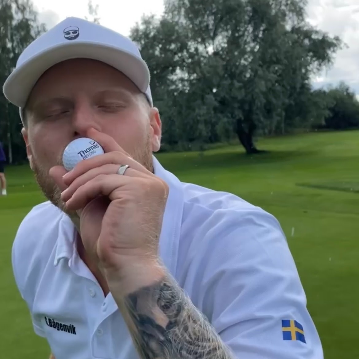 Man med tatuerad arm kysser en golfboll med texten "Thomas", iförd vit keps och tröja med svensk flagga på ärmen på golfbana.