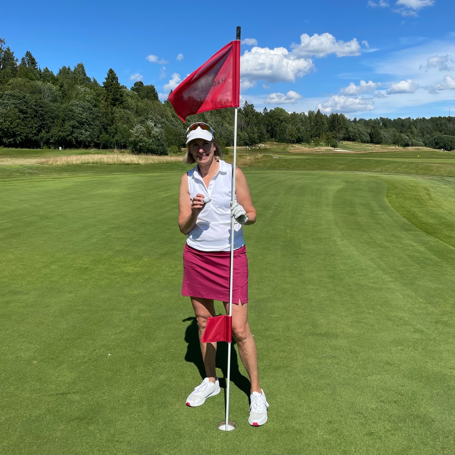 Kvinde i golfbeklædning holder en golfbold og flagstang på greenen på en solrig dag med blå himmel og skov i baggrunden.