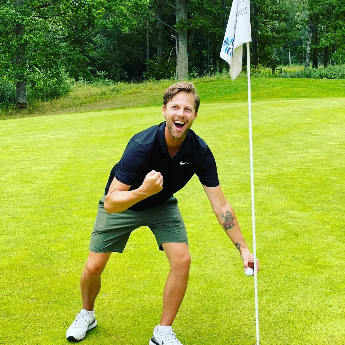 Man fejrer ved golfhul, holder golfbold og flagstang, smiler stort på grøn golfbane omgivet af træer.