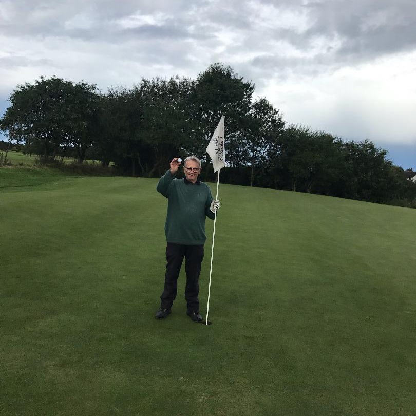 En mand står på en golfbane, holder en golfbold i den ene hånd og flagstangen i den anden, med træer i baggrunden.