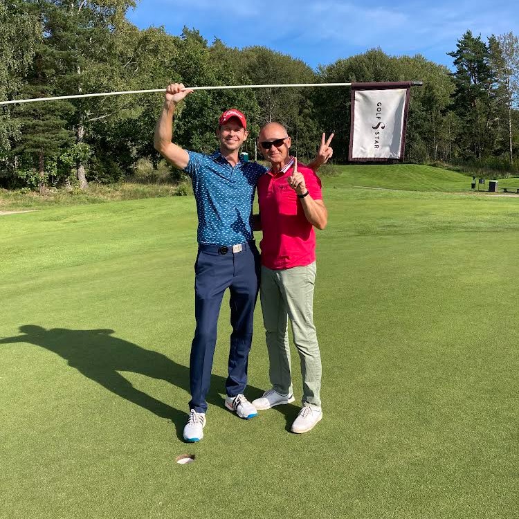 Två män står på en golfbana, en håller i en flaggstång med flagga där det står "GolfStar", båda gör segertecken.