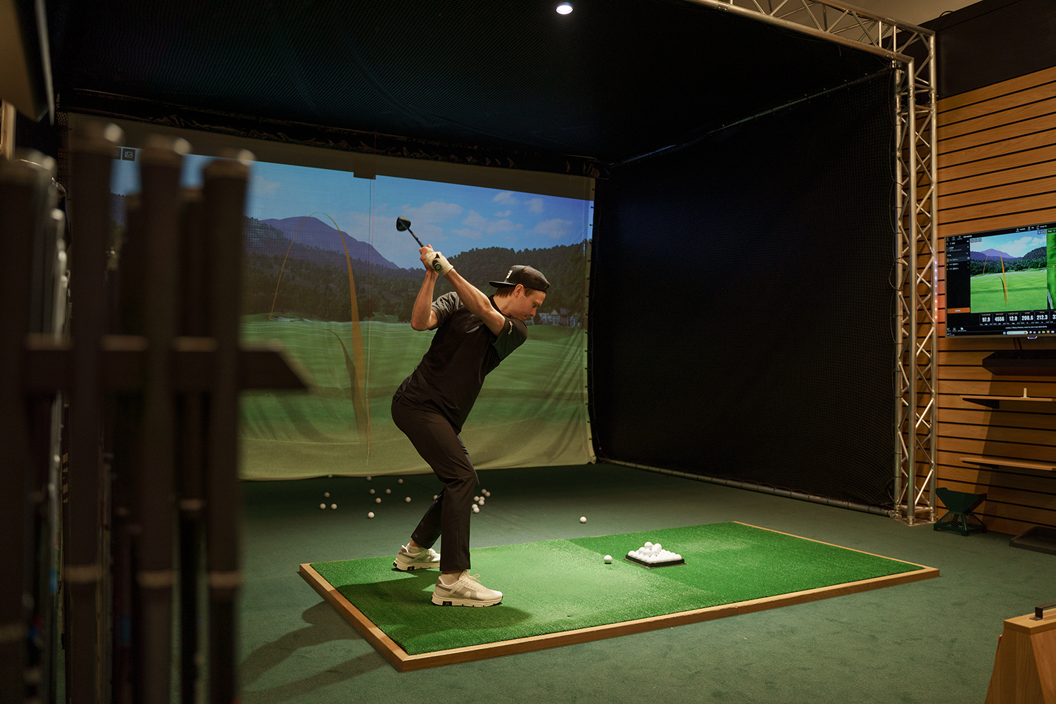 En person svingar en golfklubba på en inomhusgolfbana med golfsimulator och konstgräsmatta.