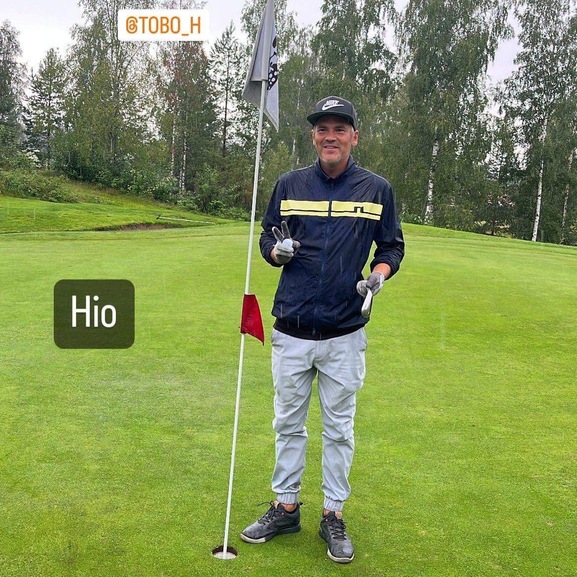 Man i golfkläder står bredvid ett hål med flagga på en golfbana, håller upp två fingrar och ler, texten "Hio" och "@TOBO_H" syns.