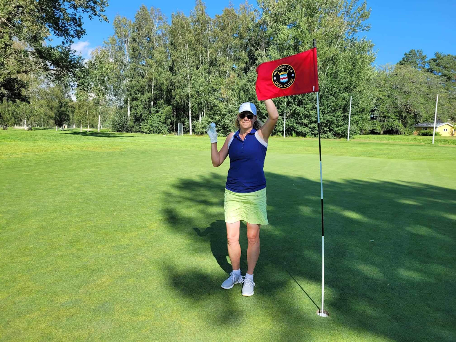 Kvinde i golfbeklædning fejrer ved flagstang med rød flag, som har teksten "Örnsköldsviks Golfklubb" på en grøn golfbane.