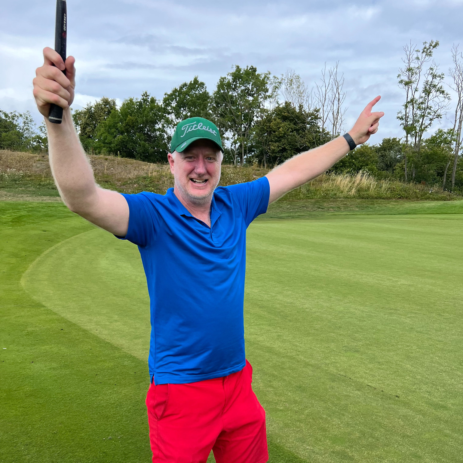 Mand i blå trøje og røde shorts fejrer med armene løftet på en golfbane, holder en golfkølle og smiler.