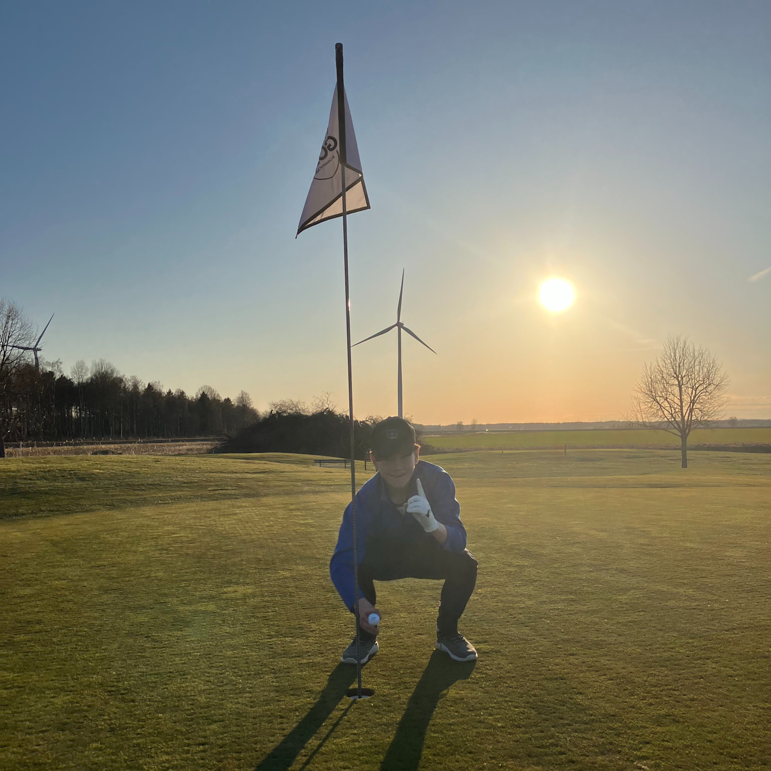 Person hukker ved et golfhul og holder en golfbold ved siden af flagstangen på en golfbane i solnedgang, med vindmøller i baggrunden.