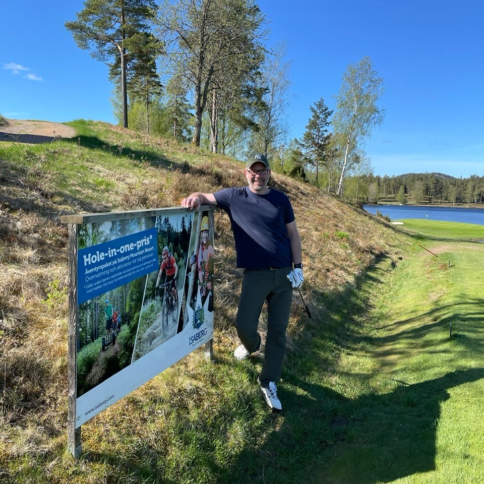 Mand iført golfbeklædning læner sig op ad et skilt, der annoncerer "Hole-in-one-præmie" ved en golfbane med en sø og træer i baggrunden.
