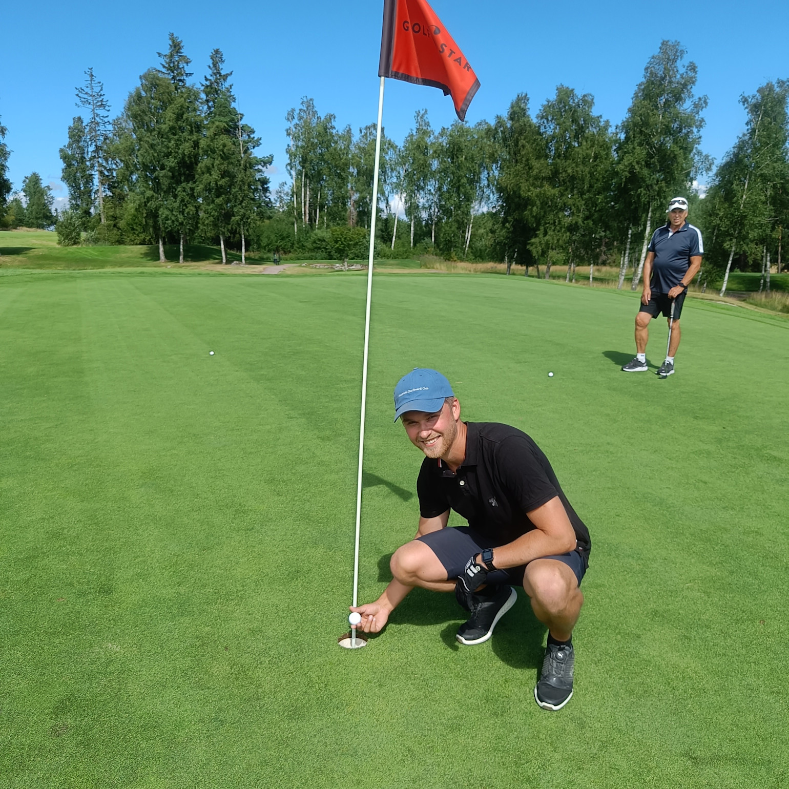 Mand i blå kasket sætter sig på hug ved golfhullet og holder en golfbold, med en anden golfspiller i baggrunden på greenen.