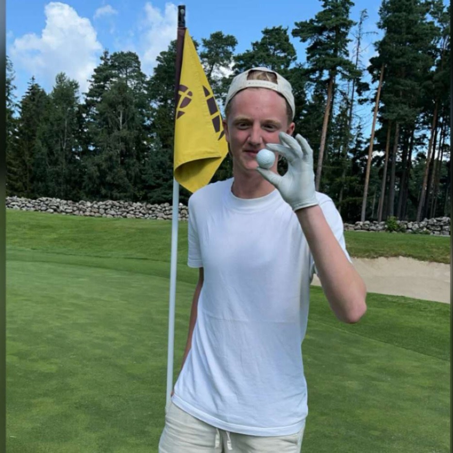Ung man i vit t-shirt och keps håller upp en golfboll framför hålet på en golfbana med flagga och träd i bakgrunden.