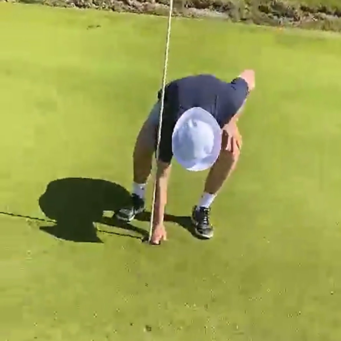 A person bends down to pick up a golf ball by the hole on a golf course in sunny weather.