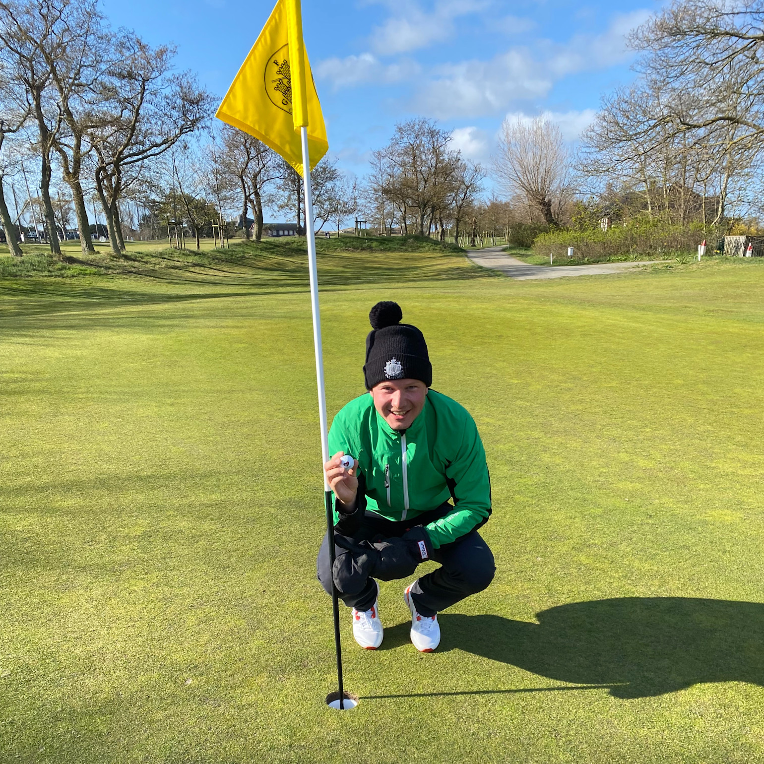 Person i grön jacka och svart mössa hukar vid en golfhål med flagga, håller en golfboll på en solig golfbana.