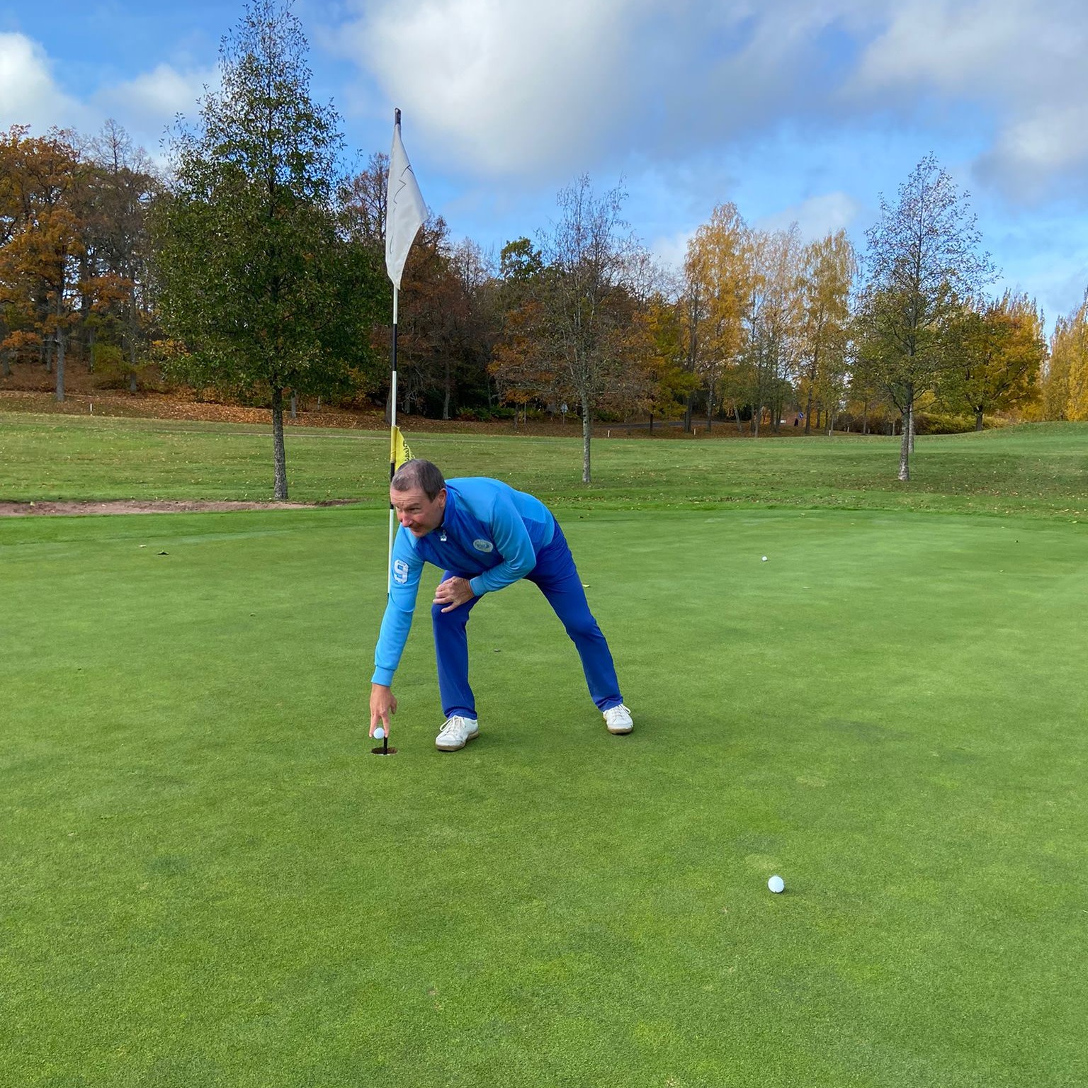 Mand i blåt golf­tøj samler en golf­bold op af hullet på greenen, med flagstang og efterårstræer i baggrunden.