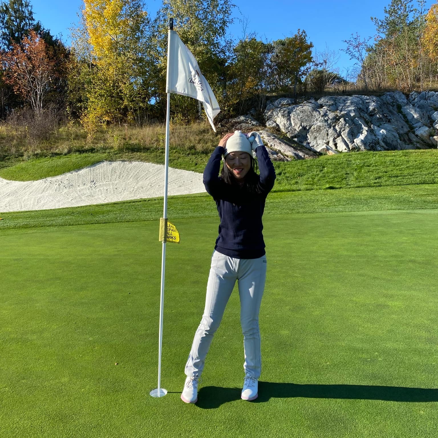 Kvinna står leende bredvid en golfflagga på en grön golfbana, omgiven av höstträd och klippor under klarblå himmel.