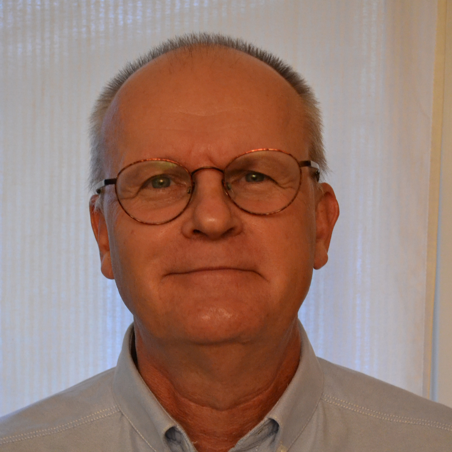 Äldre man med glasögon och kort grått hår tittar rakt fram, bär en ljusblå skjorta, neutral bakgrund.