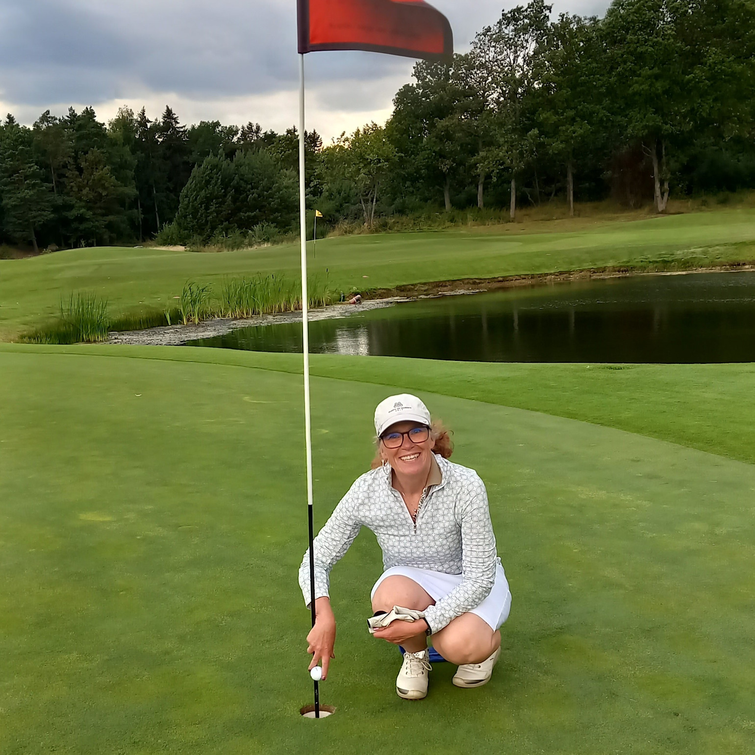 Kvinde iført golfbeklædning sidder på hug ved hullet på en golfbane, holder en golfbold og smiler, med sandbunker og træer i baggrunden.